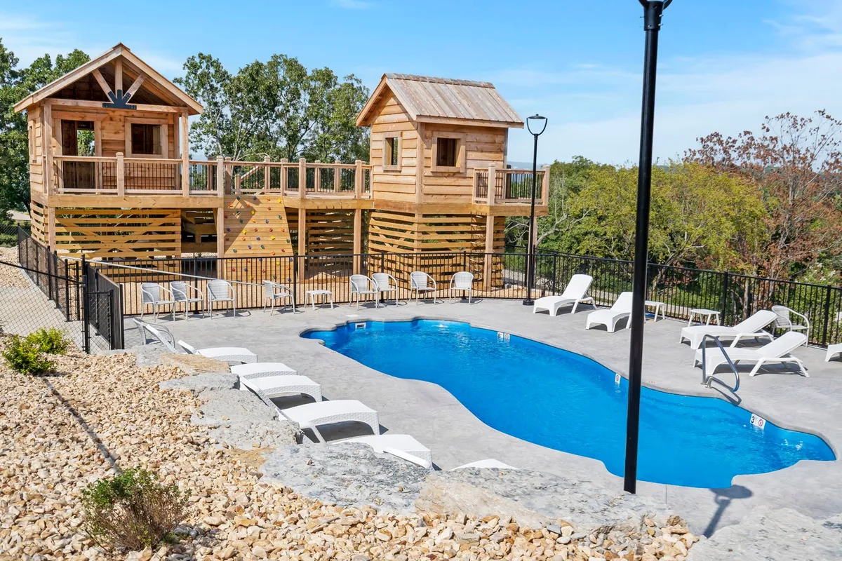 This exterior image showcases a sparkling pool surrounded by comfortable lounge chairs and playful treehouses in the background. The area is framed by greenery and clear skies, inviting both relaxation and family fun.
