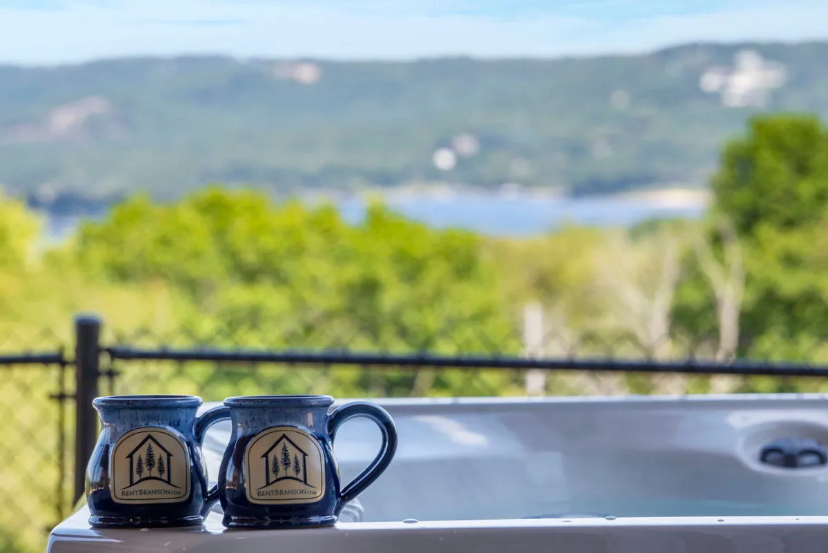 This image features two blue mugs resting on the edge of a hot tub, overlooking a serene scene of rolling hills and Table Rock Lake. The foreground suggests someone is enjoying morning coffee or relaxing in the hot tub, while the background captures the expansive water and lush landscape.