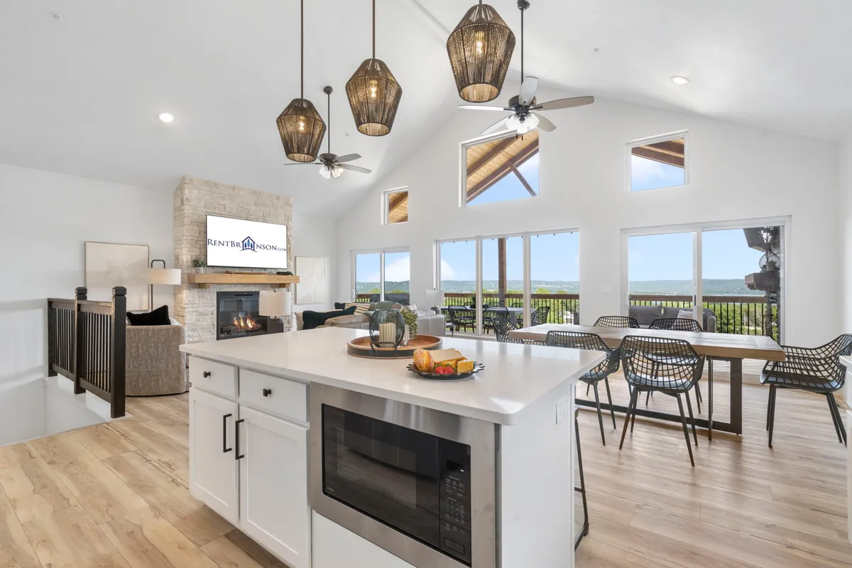 This image captures a sunlit open-concept area with a kitchen island topped with a fruit bowl, hanging pendant lights, a stone fireplace, and large windows that reveal panoramic lake views. The scene feels lively and warm, ideal for gatherings and daily living.