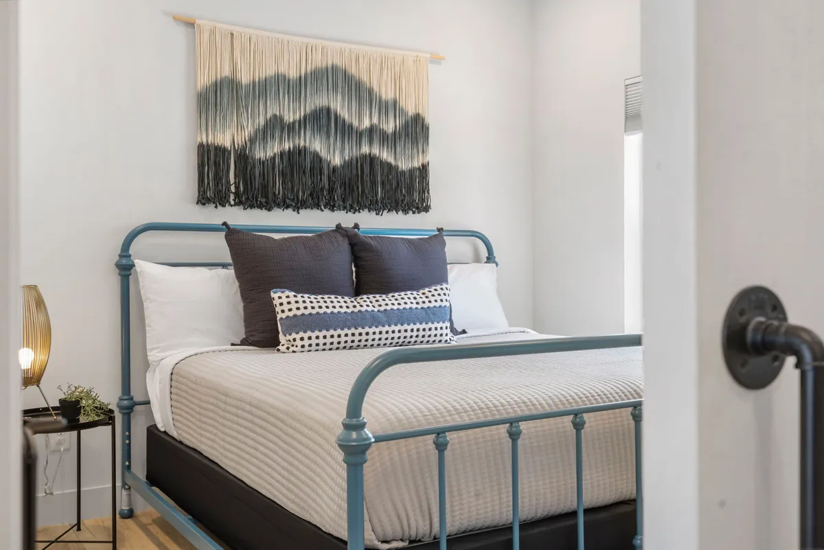 A cozy bedroom with a blue metal bed frame, decorative wall hanging, polka-dot pillows, and a nightstand with a lamp. The space is filled with soft, natural light, creating a serene and inviting atmosphere.