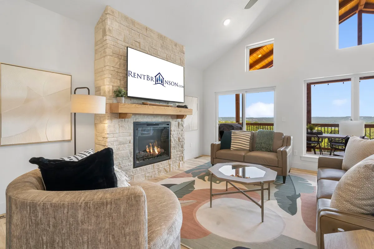 A cozy living room scene with a stone fireplace, television, large windows looking out to scenic views, and plush seating arranged for relaxation or socializing. Warm light fills the room, suggesting comfort and togetherness.