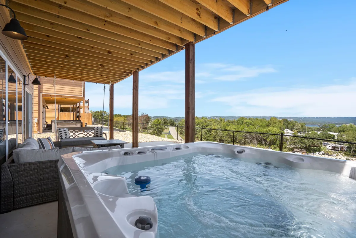 A private hot tub sits on a serene lower-level patio, surrounded by comfortable seating and framed by stunning views of the landscape and lake. The mood is tranquil, suggesting a perfect spot for soaking, relaxing, or enjoying outdoor meals against a scenic backdrop.