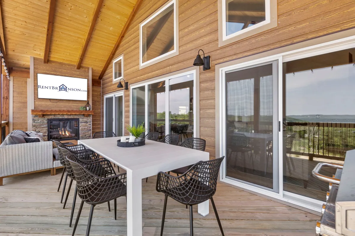 An outdoor dining area featuring a large table, stone fireplace, and comfortable seating with a scenic view of the landscape. The setting is warm and inviting, ideal for group meals, socializing, and relaxing outdoors.