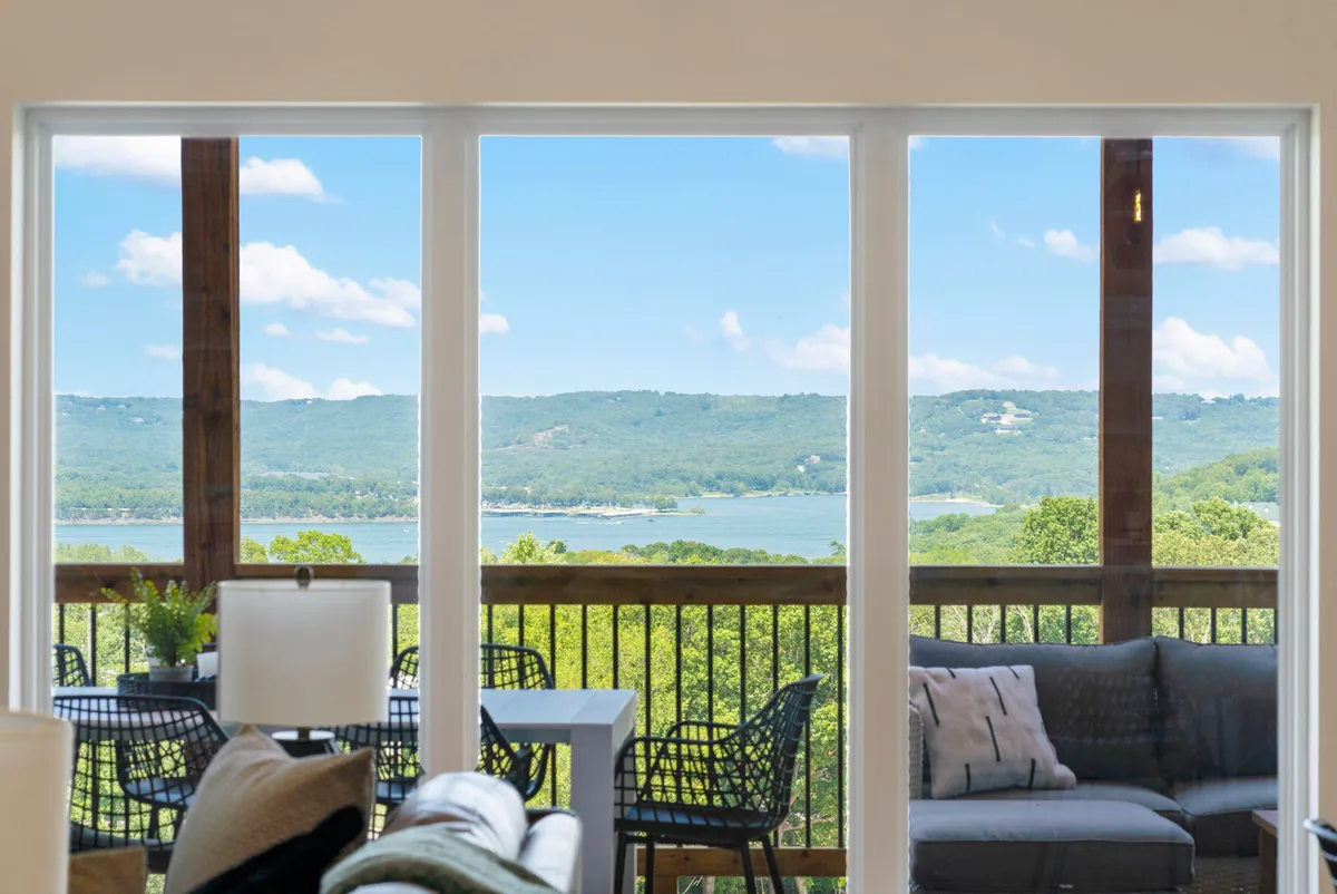 The image showcases expansive floor-to-ceiling windows framing a breathtaking, panoramic view of Table Rock Lake and rolling green hills. An inviting outdoor seating area is set up on the deck, perfect for relaxing or dining while soaking in the tranquil lake scenery. The calm mood suggests it’s an ideal spot for unwinding, entertaining, and savoring the beauty of each sunrise or sunset.